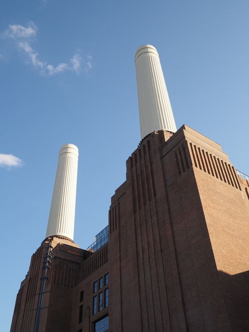 A low-angle view of the iconic Battersea Power Station with its large brick facade and four distinctive white chimneys rising against a clear blue sky. The brick exterior shows well-maintained surfaces, with visible vertical ridges and rectangular grid patterns around the structure. Sunlight illuminates the building, highlighting the clean and preserved appearance of the historic industrial architecture. The photograph captures the grandeur of the power station, emphasizing its architectural details and prominent chimney stacks, set in an urban environment. Battersea Carpet Cleaning’s logo or branding is not visible, but the image relates to the original page about surface cleaning, deep cleaning, and sanitisation services in Battersea.