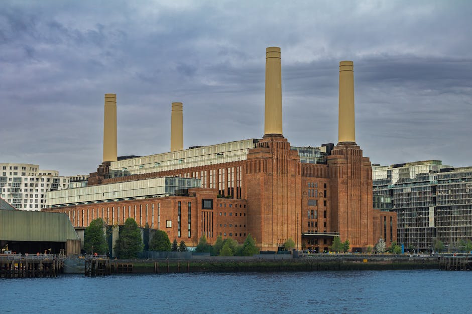 A low-angle view of the iconic Battersea Power Station with its large brick facade and four distinctive white chimneys rising against a clear blue sky. The brick exterior shows well-maintained surfaces, with visible vertical ridges and rectangular grid patterns around the structure. Sunlight illuminates the building, highlighting the clean and preserved appearance of the historic industrial architecture. The photograph captures the grandeur of the power station, emphasizing its architectural details and prominent chimney stacks, set in an urban environment. Battersea Carpet Cleaning’s logo or branding is not visible, but the image relates to the original page about surface cleaning, deep cleaning, and sanitisation services in Battersea.