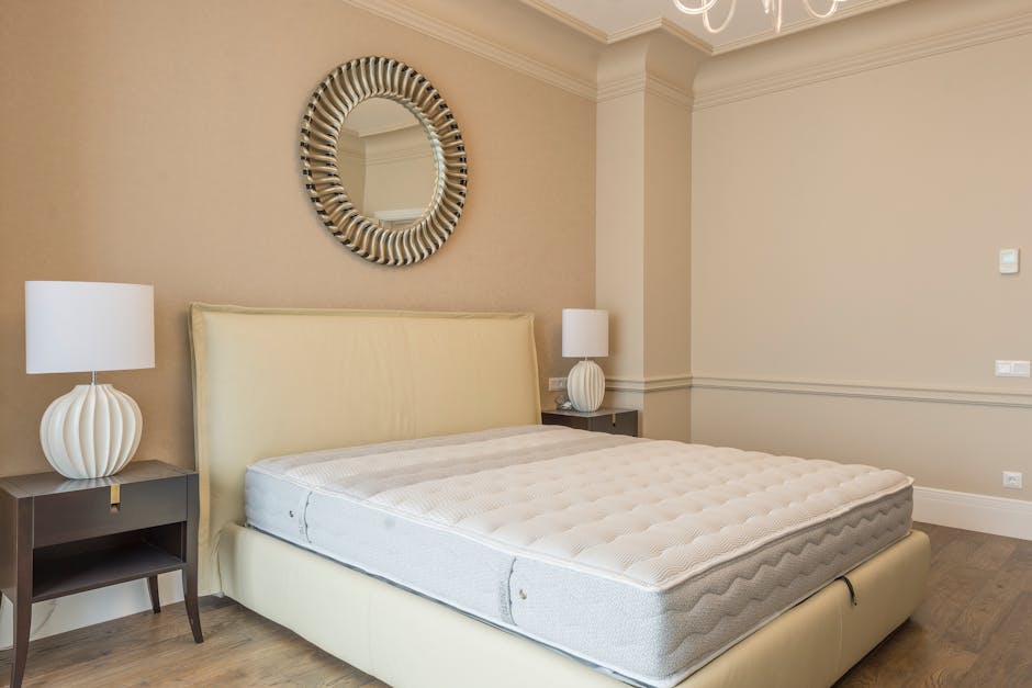 A modern bedroom featuring a cream upholstered bed with a newly cleaned white mattress, flanked by two dark wood bedside tables, each topped with a matching white ceramic table lamp with rounded bases. The room has beige walls with crown moulding and a circular wall mirror with a decorative silver frame hanging above the headboard. The floors are light wood, and the space appears tidy and well-maintained, reflecting professional domestic surface cleaning and hygiene practices in line with Battersea Carpet Cleaning's mattress cleaning and stain removal guide.