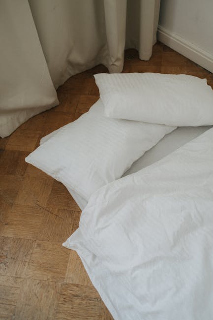 A close-up view of a domestic scene on a wooden floor, featuring a bed with crisp white bedding and pillows. The bedding appears clean and neatly arranged, with the pillows placed on the floor next to the bed. In the background, light-colored curtains partially cover a window, and the room is lit with natural light, highlighting the smooth, polished surface of the wooden floor. The overall impression is of a tidy, hygienic environment suitable for surface cleaning, deep cleaning, or sanitisation, as part of routine domestic cleaning. Battersea Carpet Cleaning offers professional cleaning services which include maintaining cleanliness and hygiene in residential spaces.