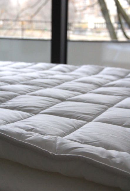 A modern bedroom featuring a cream upholstered bed with a newly cleaned white mattress, flanked by two dark wood bedside tables, each topped with a matching white ceramic table lamp with rounded bases. The room has beige walls with crown moulding and a circular wall mirror with a decorative silver frame hanging above the headboard. The floors are light wood, and the space appears tidy and well-maintained, reflecting professional domestic surface cleaning and hygiene practices in line with Battersea Carpet Cleaning's mattress cleaning and stain removal guide.