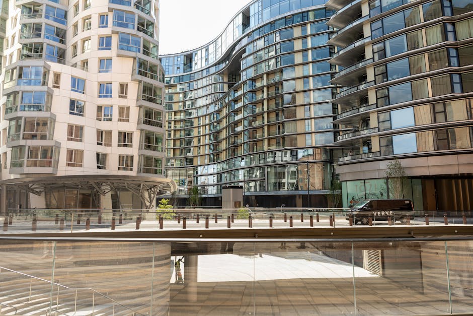 Exterior view of modern high-rise residential buildings with curved glass facades and numerous balconies, featuring a mix of white and dark grey materials. The ground level includes a paved area with metal bollards and a glass railing. Natural daylight illuminates the scene, highlighting the clean, reflective surfaces of the buildings. Battersea Carpet Cleaning offers surface cleaning and deep cleaning services, ensuring hygiene and presentation for both residential and commercial properties in Battersea, as showcased by this contemporary urban setting.
