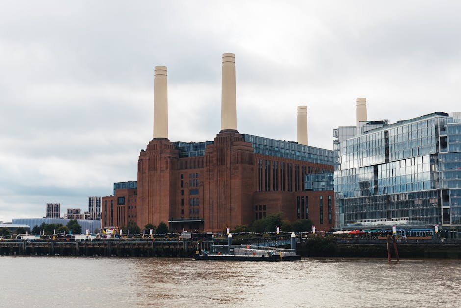 A large industrial building, resembling a historic power station, constructed from reddish-brown brickwork, features three tall, cream-colored smokestacks. The structure is situated along a riverbank, with metal walkways and a boat dock in the foreground. Adjacent to the power station is a modern glass-fronted office building reflecting the cloudy sky, which illuminates the scene with diffused daylight. The surroundings include smaller buildings and trees, with no visible signs of cleanliness or maintenance activities. The image highlights the contrasting architectural styles of the old industrial and contemporary structures along the water’s edge, representative of urban regeneration areas like Battersea.